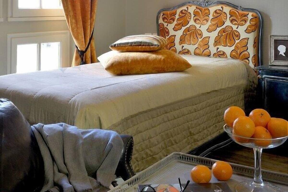 Un dormitorio con cama, una mesita de noche con una revista, un cuenco de naranjas y una ventana con cortinas.