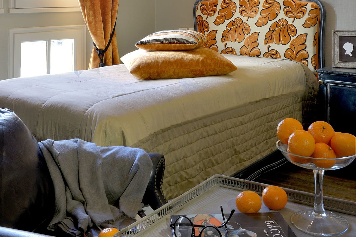 Un dormitorio con una cama, una mesita de noche con un cuenco de naranjas, una lámpara y una ventana con cortinas.