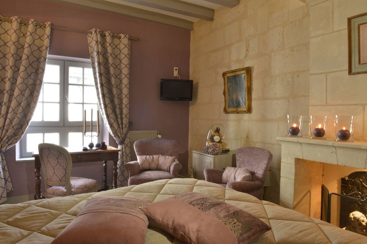 Un dormitorio con una cama grande, dos sillones, un escritorio, un televisor, una chimenea y una ventana con cortinas.