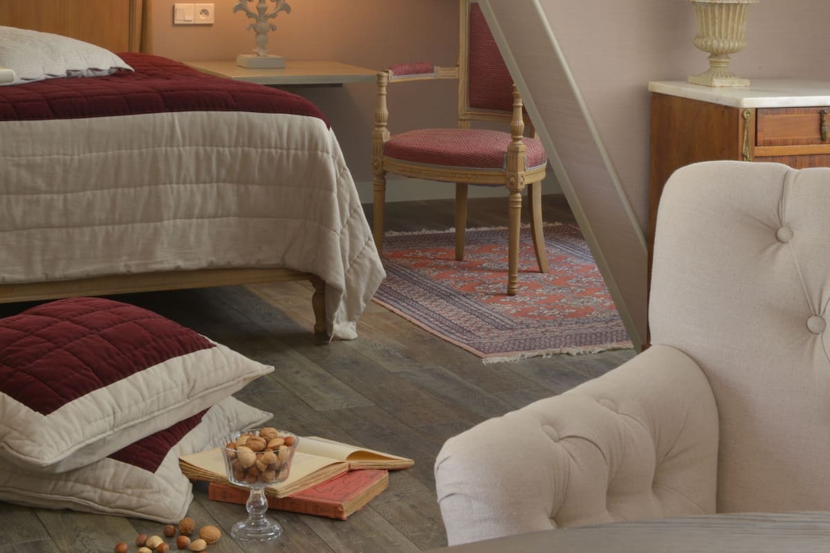 Un dormitorio con techo inclinado, una cama con edredón rojo y blanco, una mesita de noche con lámpara, una silla y una pequeña mesa con un libro y un vaso de agua.
