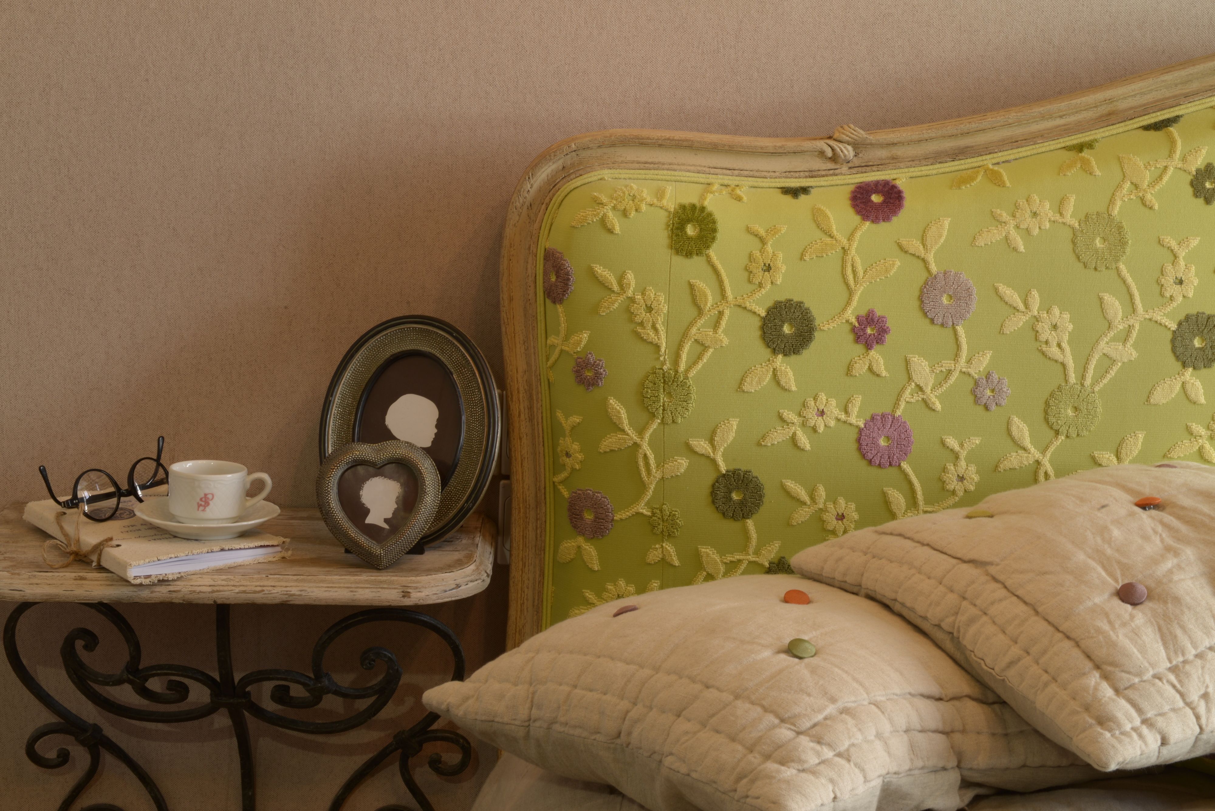 Una cama con cabecera floral, una mesita de noche con una taza, vasos y un libro, y un espejo con forma de corazón.