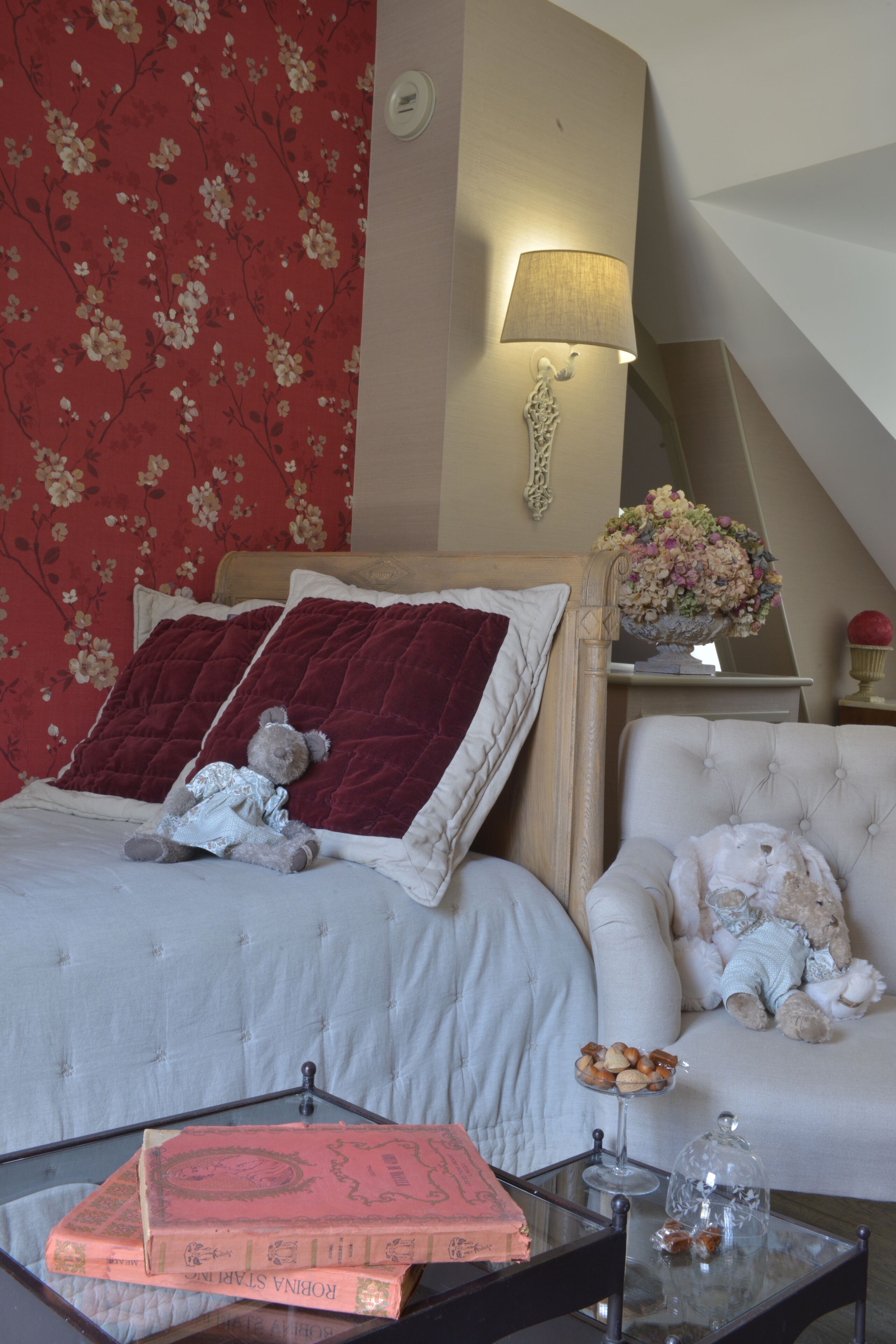 Een slaapkamer met een bed, een stoel, een lamp en een tafel met boeken en decoratieve spullen.