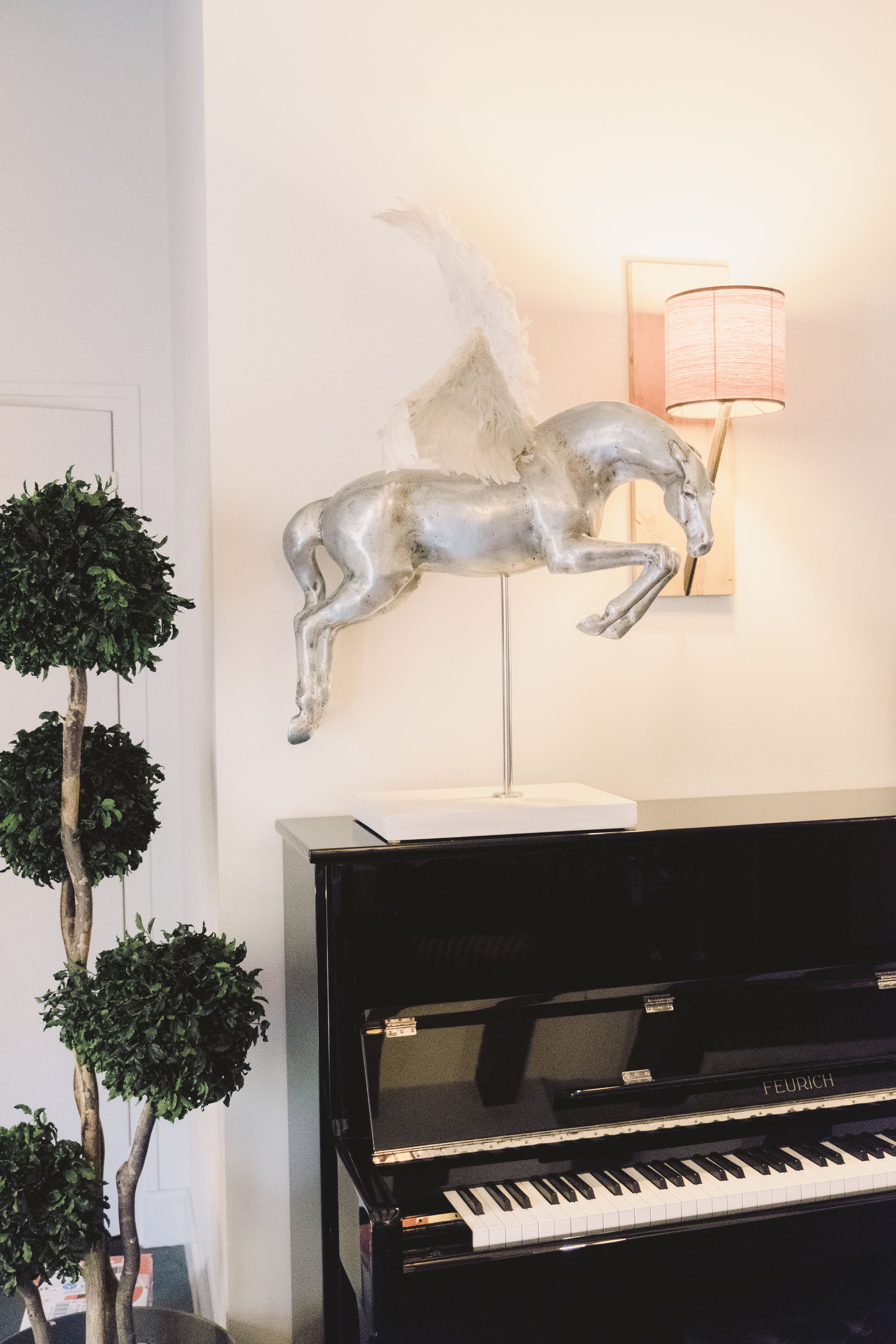 A grand piano, a wall-mounted sculpture of a horse, and a tall, slender tree.