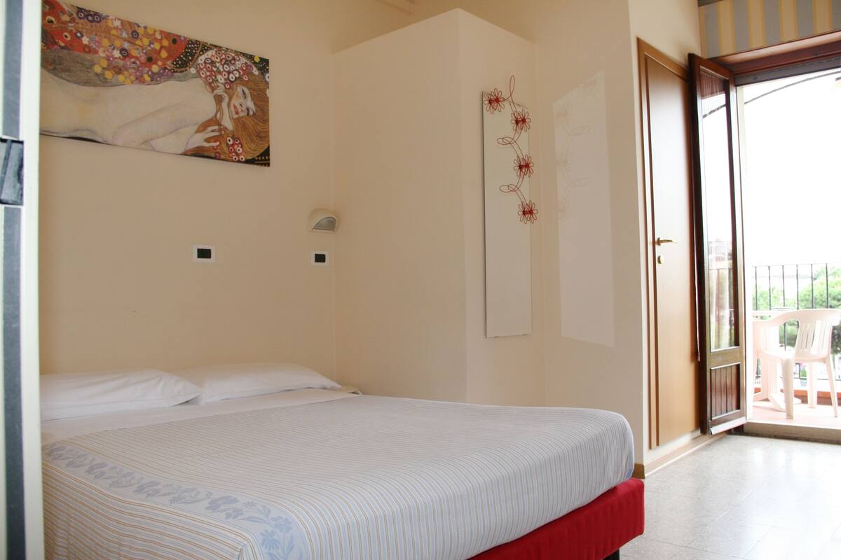 A bedroom with a bed, a painting, a mirror, and a door leading to a balcony.
