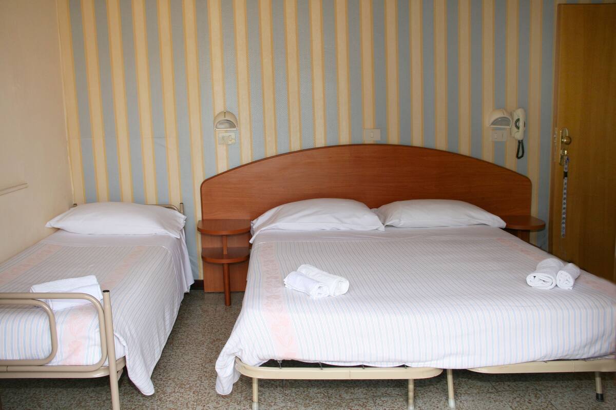A hotel room with two single beds, each with a wooden headboard, white linens, and a bedside table with a lamp.
