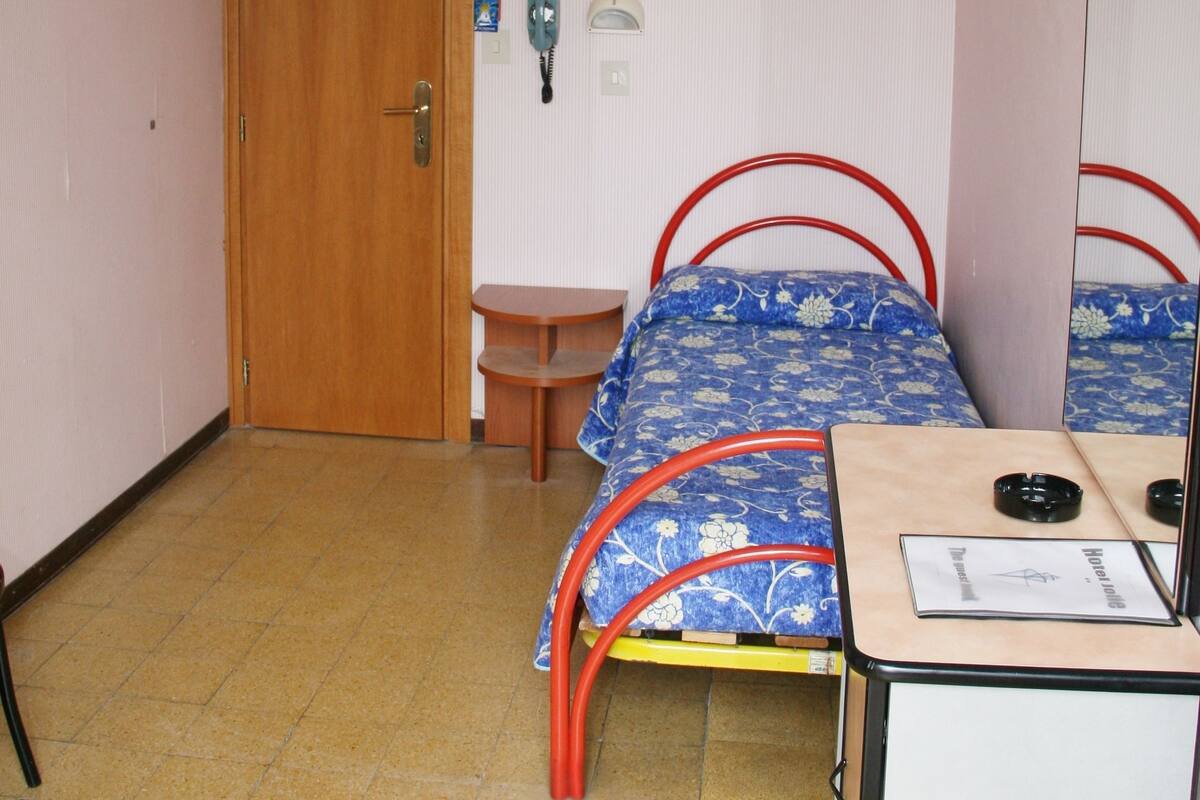 A bedroom with a wooden door, a bed with a red metal frame, a desk with a mirror, and a wall-mounted phone.
