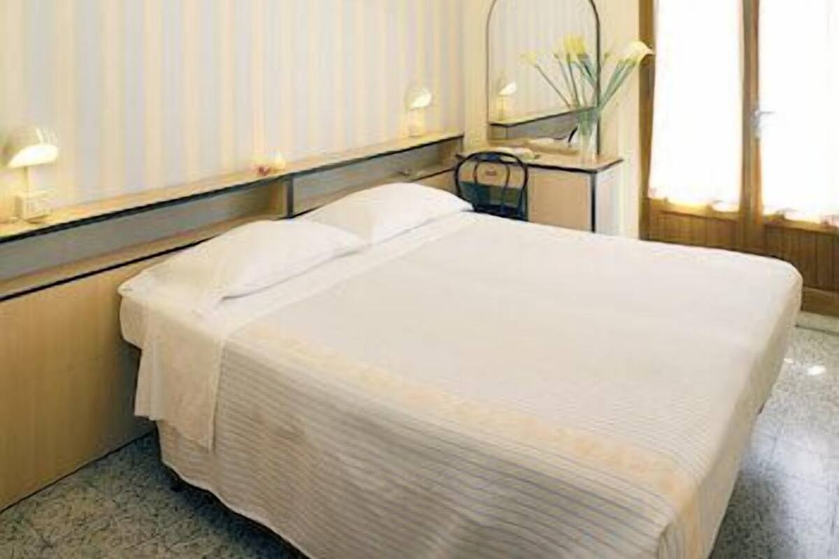 A hotel room with a bed, a nightstand with a lamp, a mirror, and a window with curtains.
