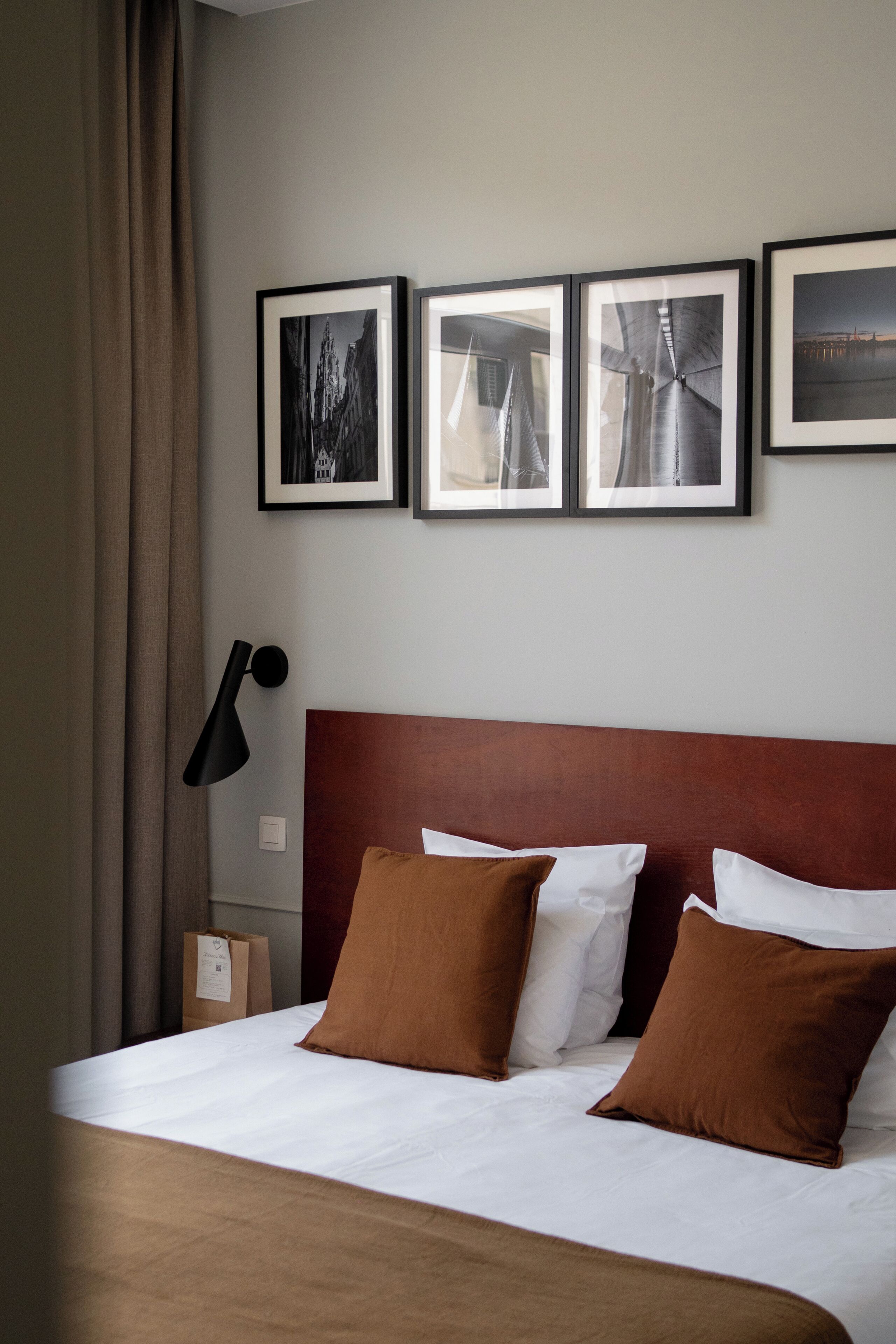 A hotel room with a bed, brown and white pillows, a headboard, a wall-mounted lamp, and three framed pictures.