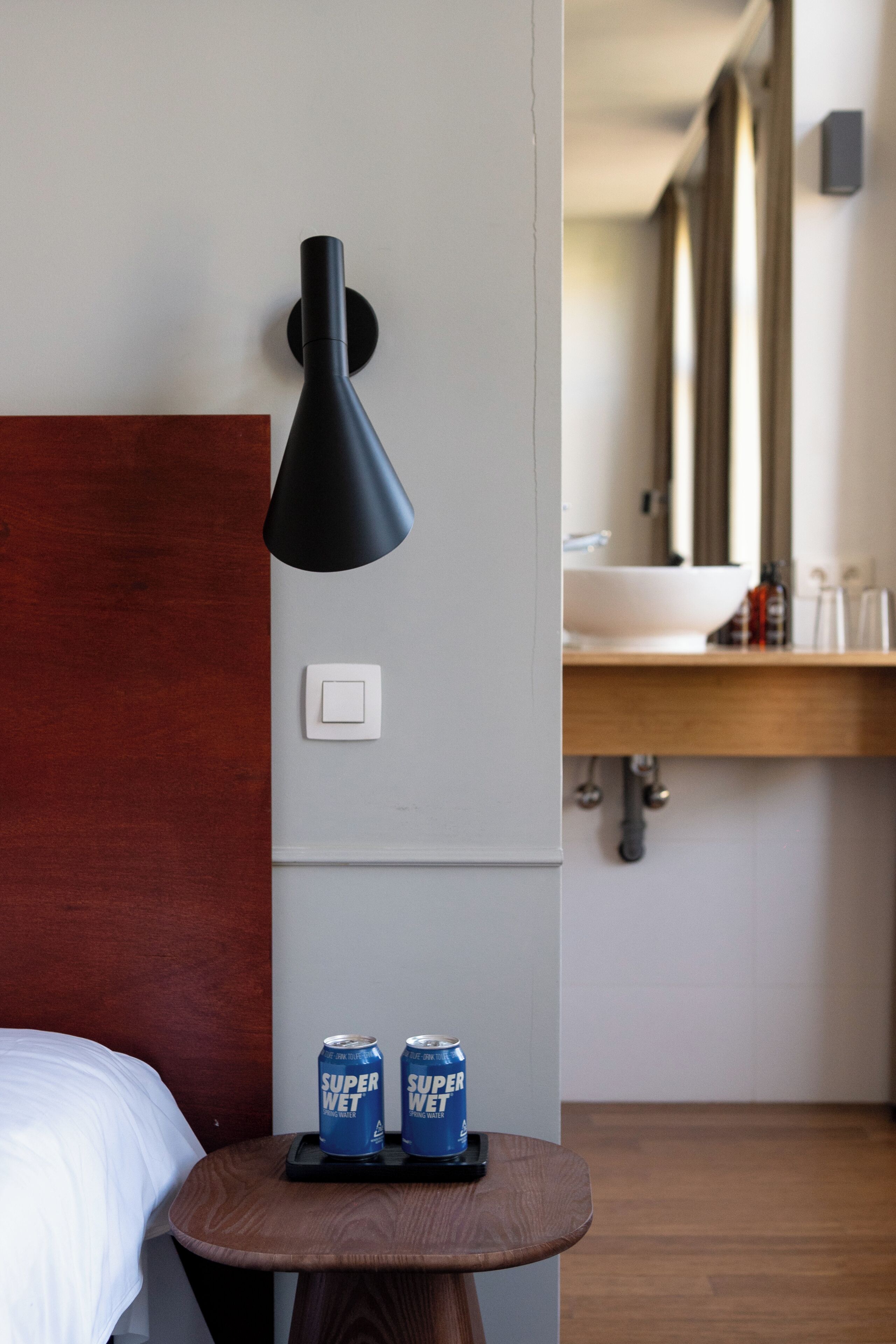 Habitación de hotel con una cama, una pequeña mesa de madera con dos latas de Super Wet, una lámpara de pared negra, un interruptor blanco y una encimera de madera con un lavabo y un espejo.