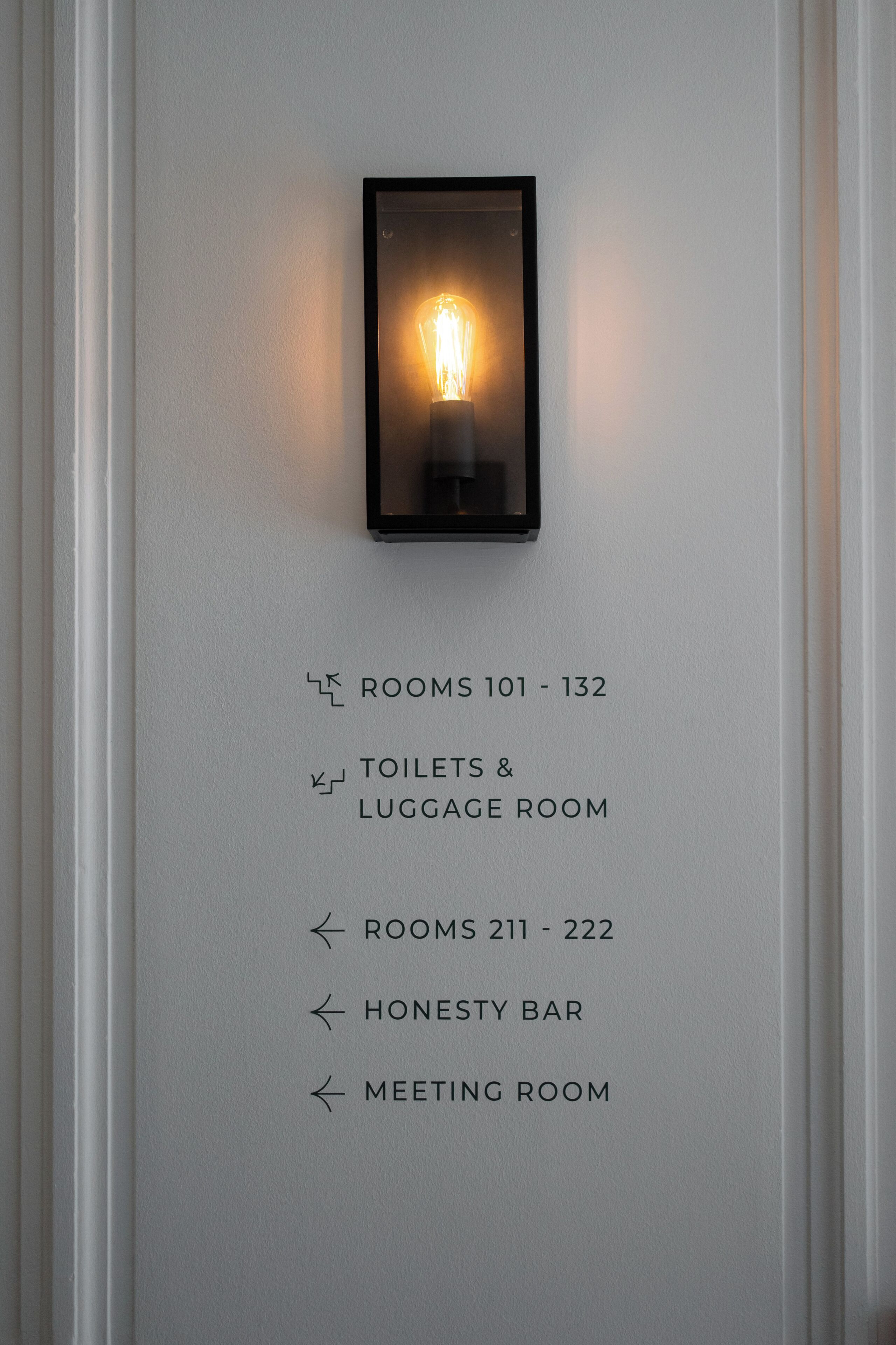 Un letrero en la pared con flechas direccionales y texto que indica números de habitaciones y servicios como baños, sala de equipaje, bar de confianza y sala de reuniones.