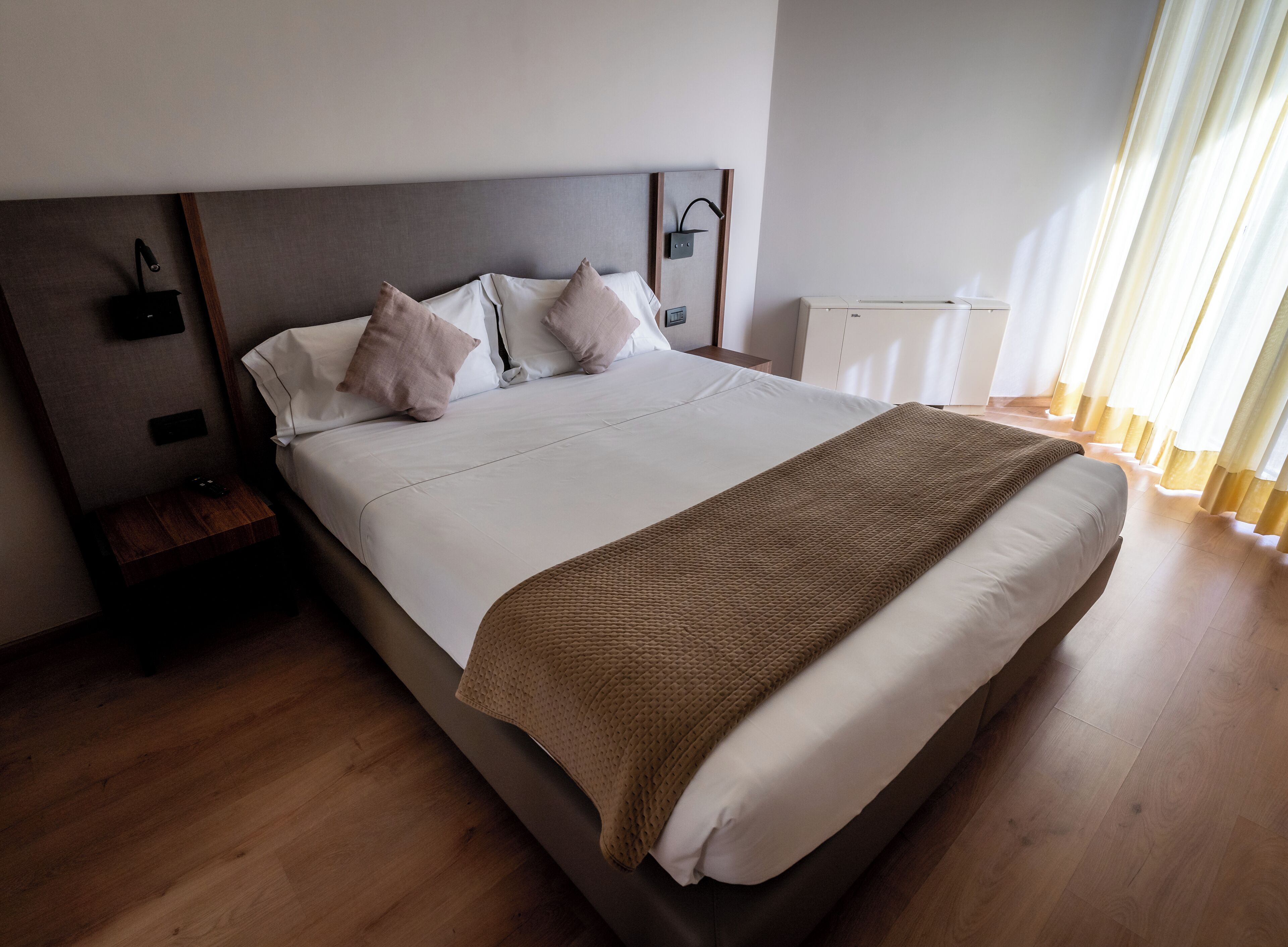 Un lit bien fait, recouvert d’un plaid marron, une table de chevet en bois avec une lampe, et une vue sur une fenêtre avec des rideaux.