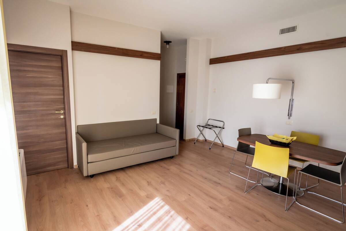 A room with a wooden floor, a sofa, a dining table with chairs, and a lamp.
