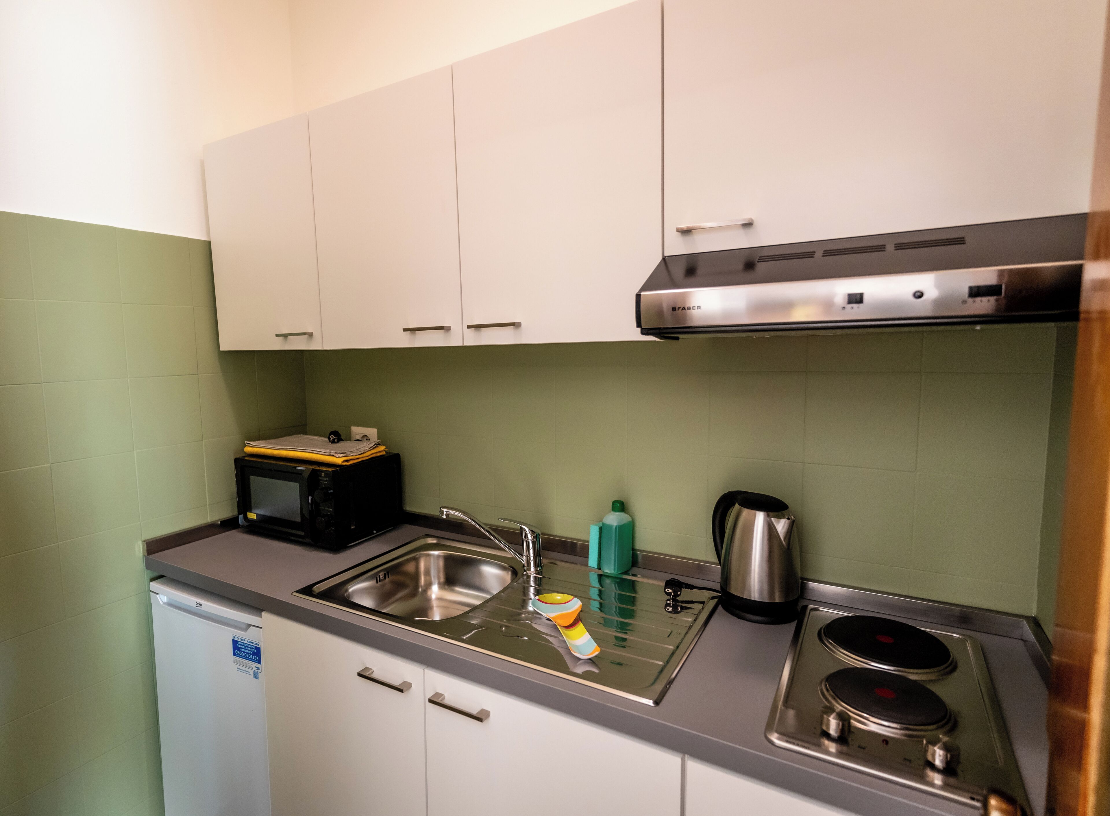 A compact kitchen with white cabinets, a stainless steel sink, a microwave, a kettle, and a stove.
