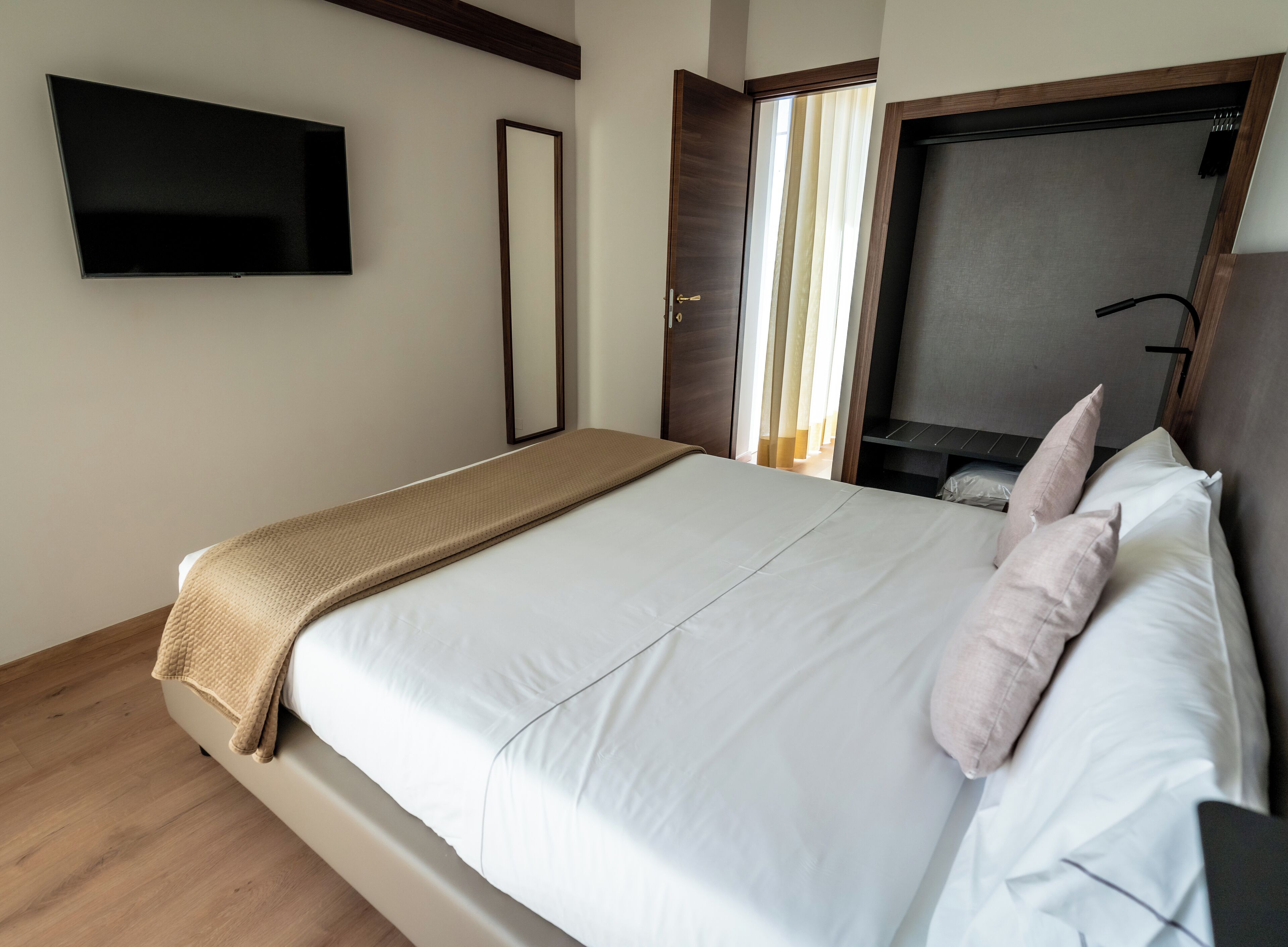 Un lit bien fait, recouvert d’un plaid beige, un téléviseur à écran plat fixé au mur et une porte en bois avec un miroir.