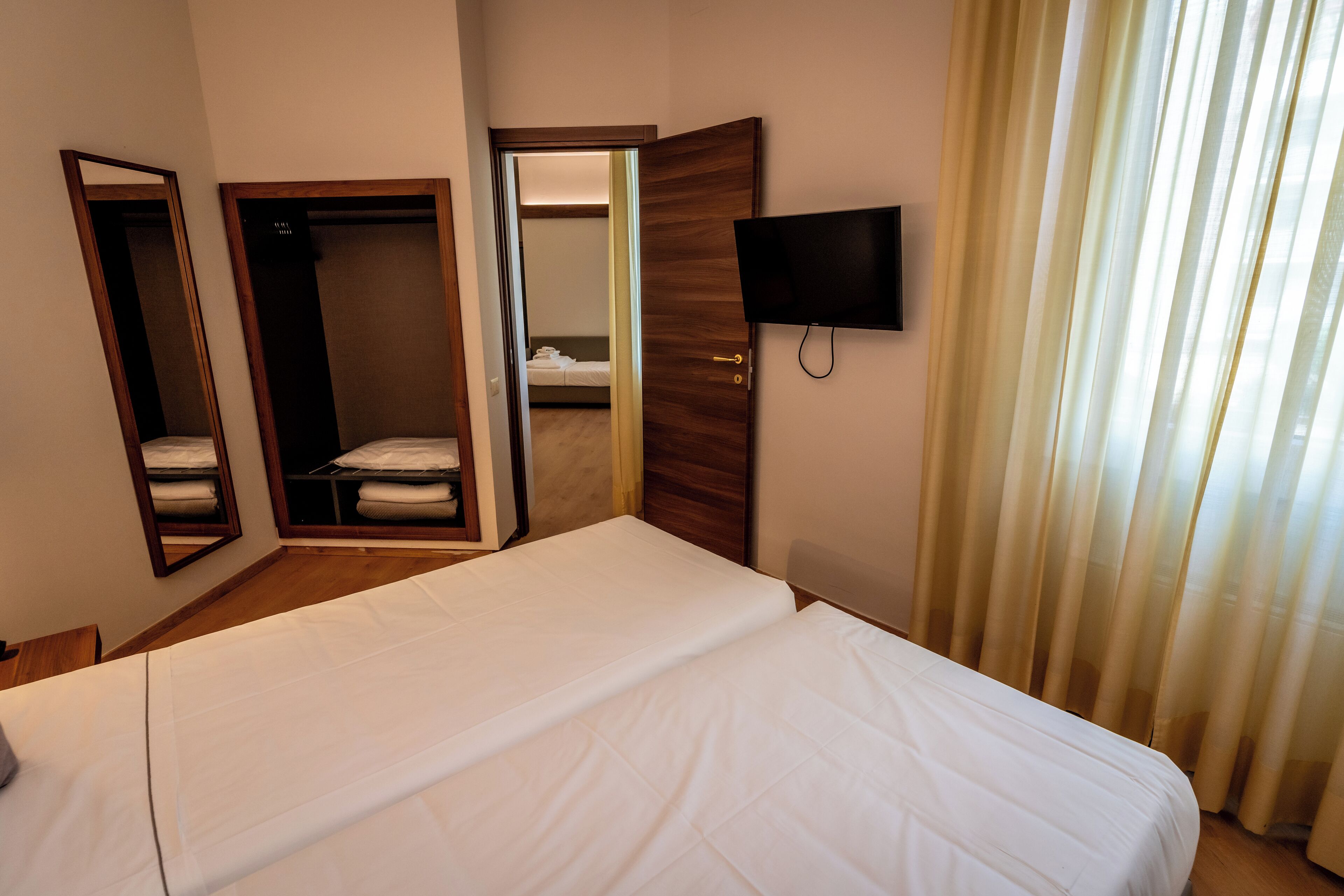 A hotel room with a bed, a wardrobe, a mirror, a television, and a window with curtains.