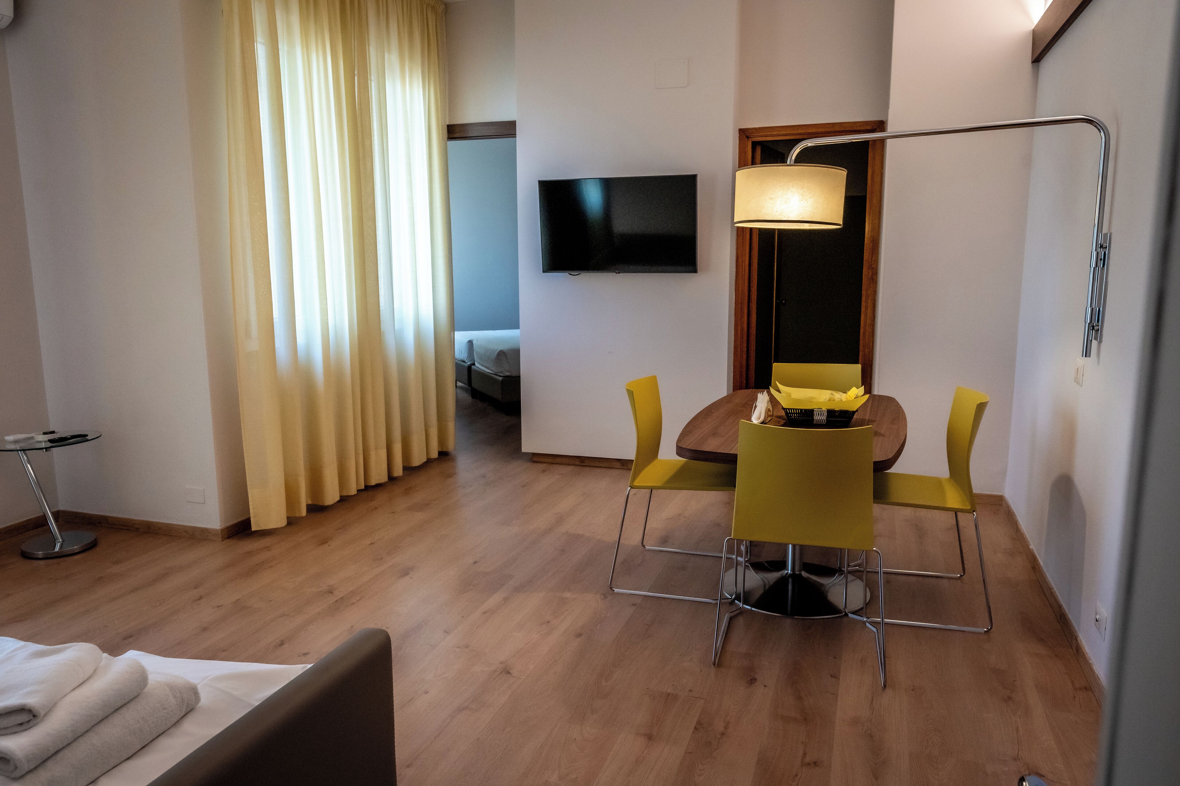 A hotel room with a bed, a desk with a chair, a TV mounted on the wall, and a window with curtains.