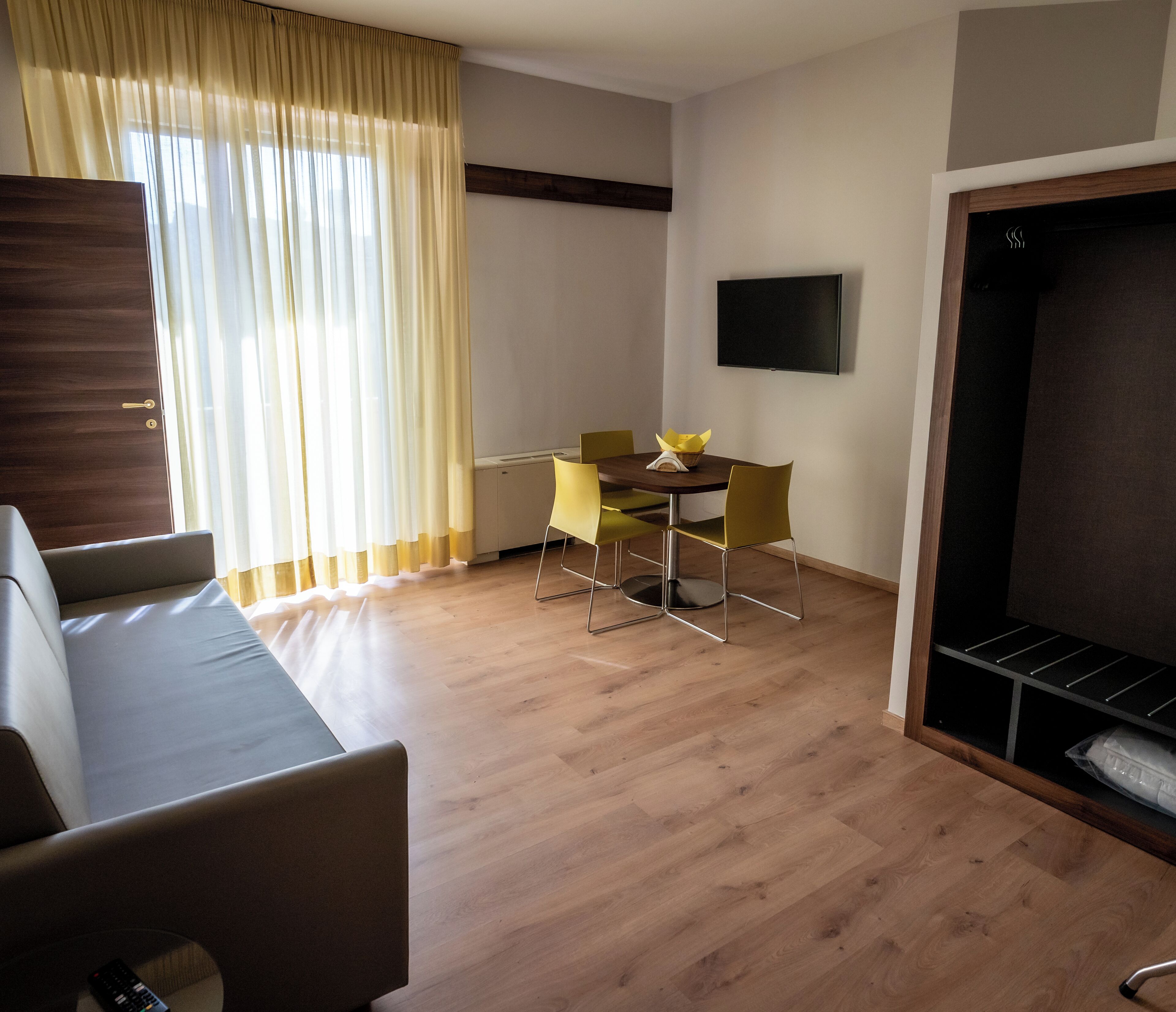 A modern living room with a wooden floor, a sofa, a dining table with yellow chairs, and a flat-screen TV mounted on the wall.