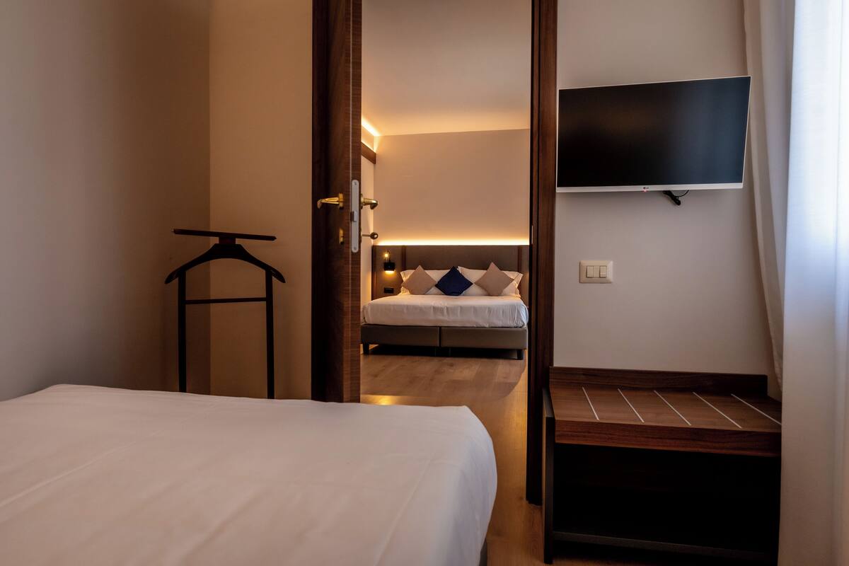 A hotel room with a bed, a hanging coat rack, a bedside table, a TV mounted on the wall, and a wooden bench.