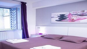 A modern bedroom with a large bed, a window with blinds, a nightstand with a lamp, and a wall-mounted artwork. - Piumith (Rome)