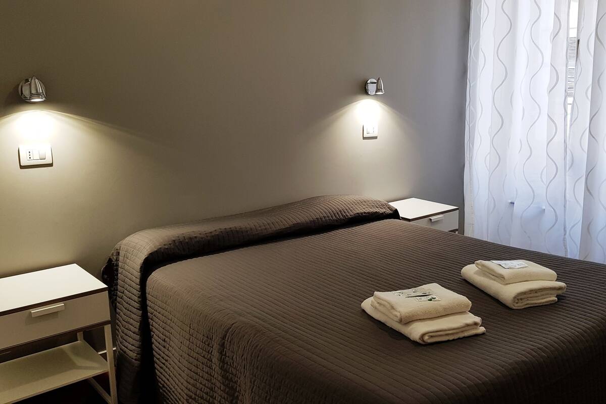 Un lit bien fait, avec deux serviettes pliées, une table de chevet, une lampe fixée au mur et une fenêtre ornée d’un rideau ondulé.