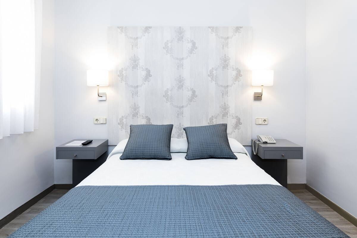 A modern hotel room with a large bed, two bedside tables, a nightstand, a telephone, and two wall-mounted lamps.