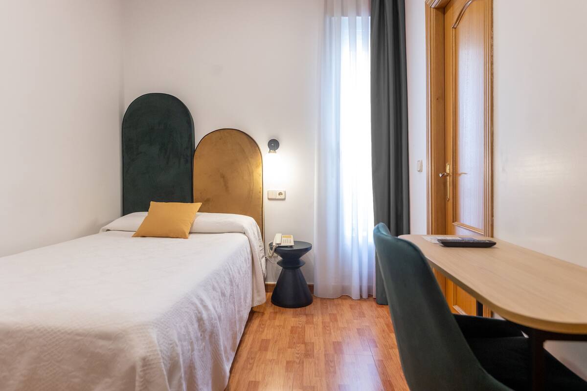 A hotel room with a bed, a desk, a chair, a telephone, and a window with curtains.