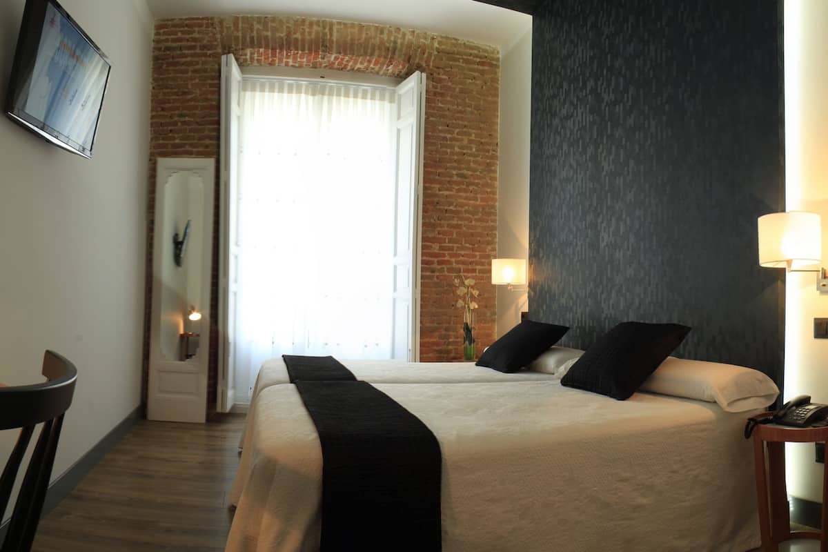 A hotel room with a bed, a desk with a lamp, a TV on the wall, and a window with curtains.