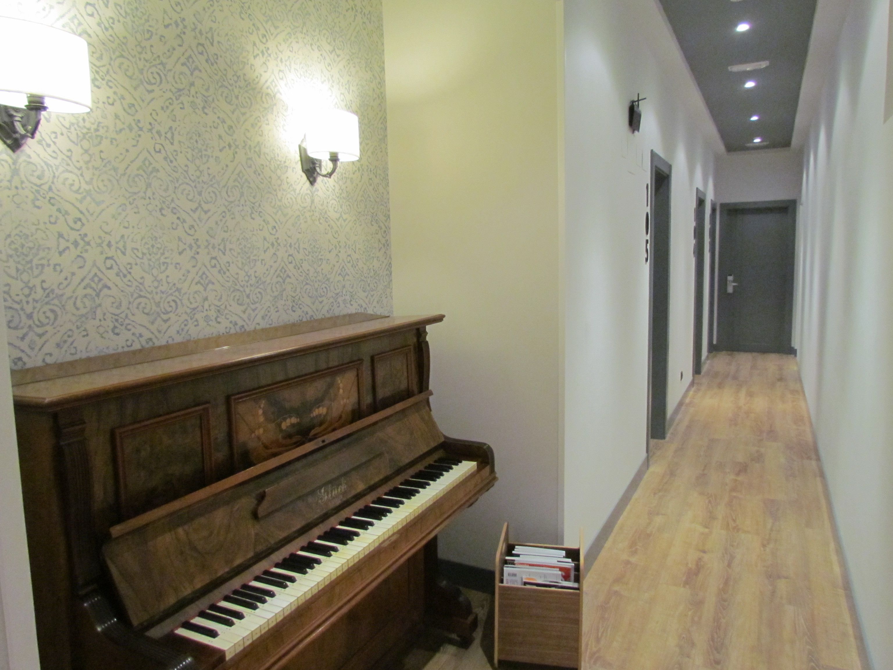 Un pasillo con suelo de madera, un piano y una lámpara de pared.