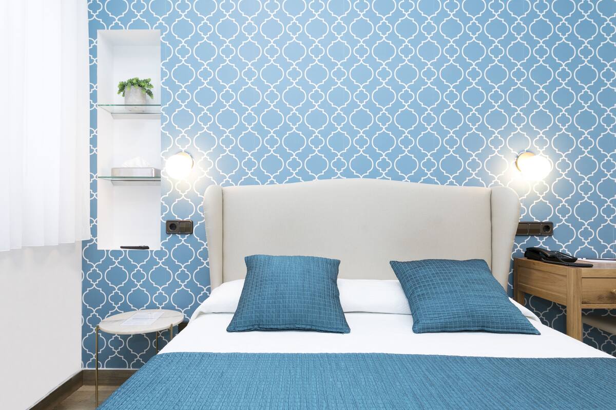 A bedroom with a bed, two pillows, a bedside table, a nightstand, a wall-mounted shelf with a plant, and a patterned wall.