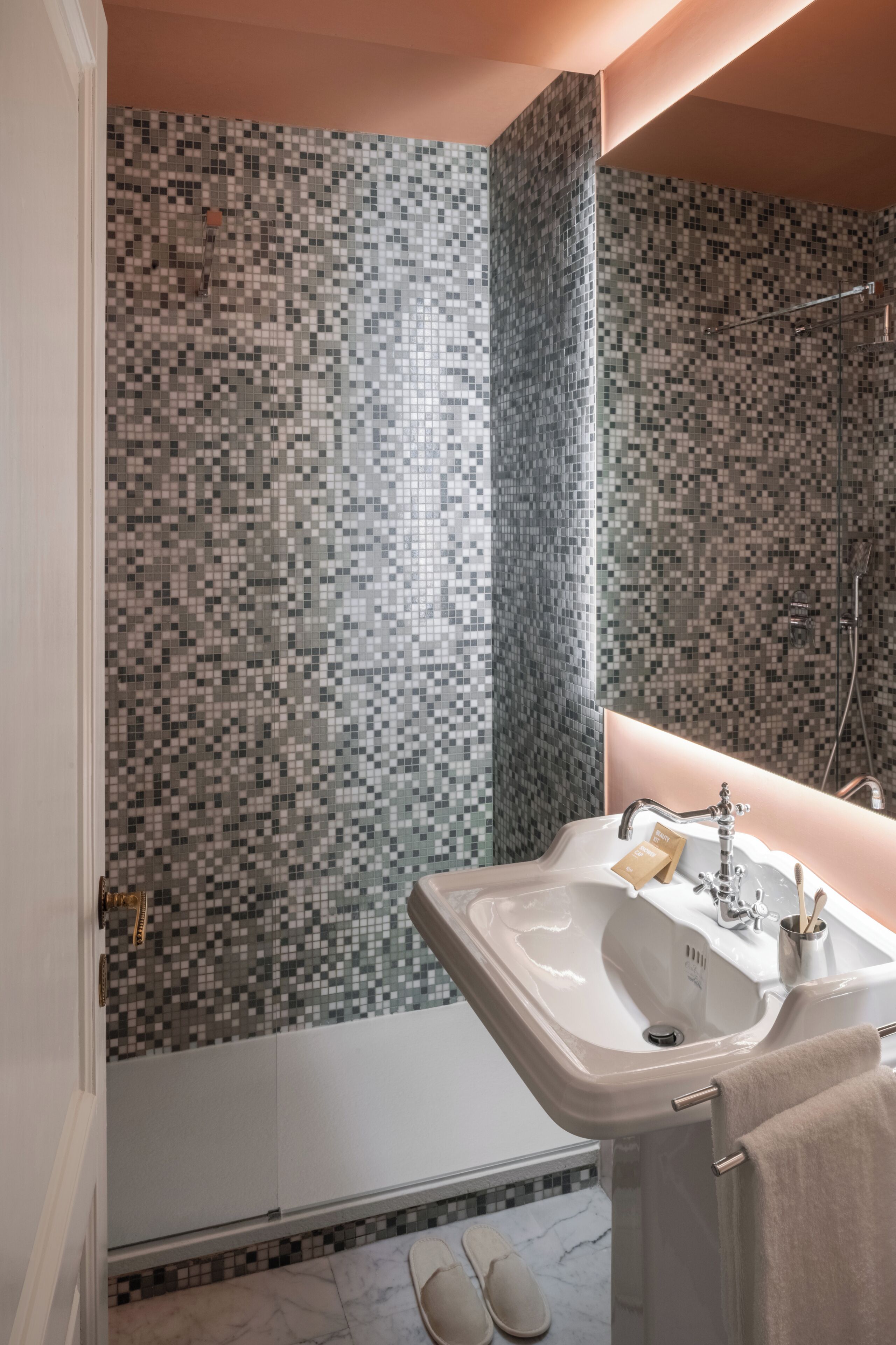 Ein Badezimmer mit gefliesten Duschwänden, einem Waschbecken mit Handtuch und einem Paar Hausschuhen auf dem Boden.