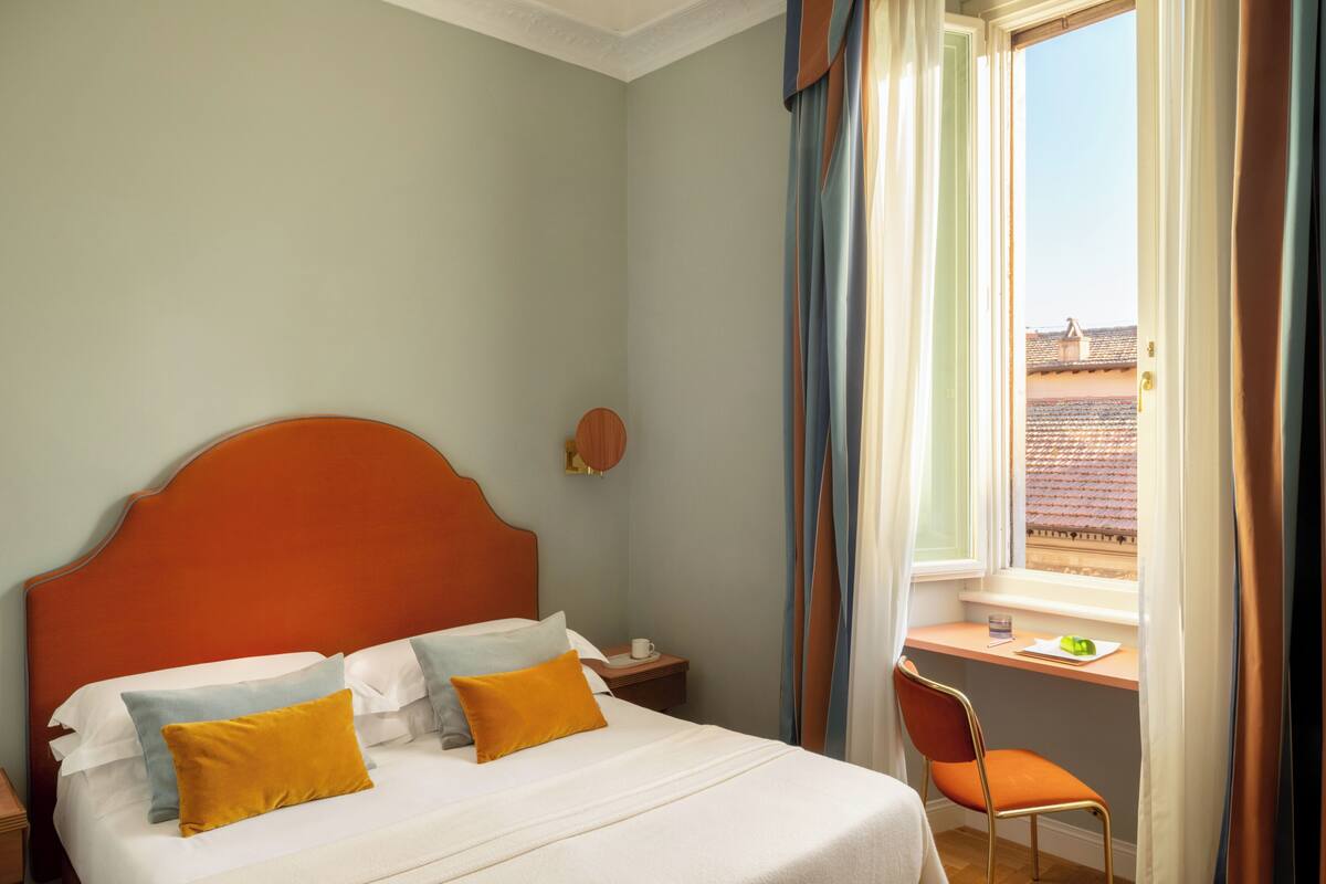 Ein Hotelzimmer mit Bett, Nachttisch, Stuhl, einem Fenster mit Vorhängen und Blick auf Gebäude.
