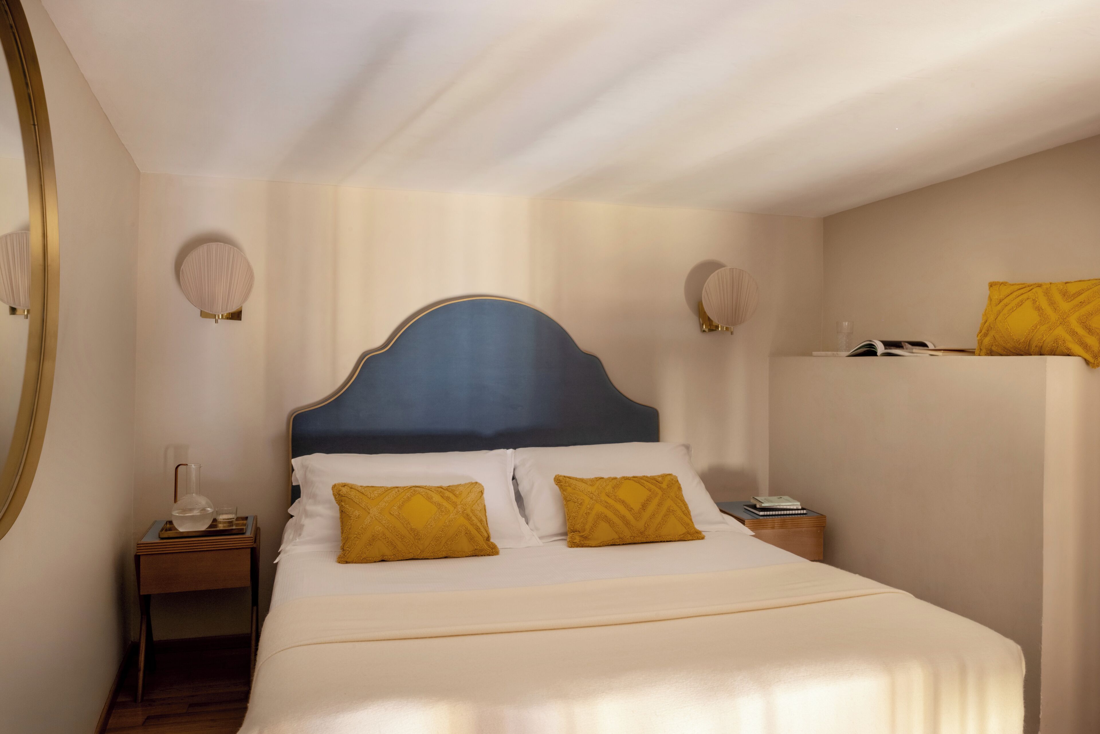 Een net opgemaakt bed met een blauw hoofdeinde, twee kussens, een nachtkastje met een lamp en een spiegel aan de muur.