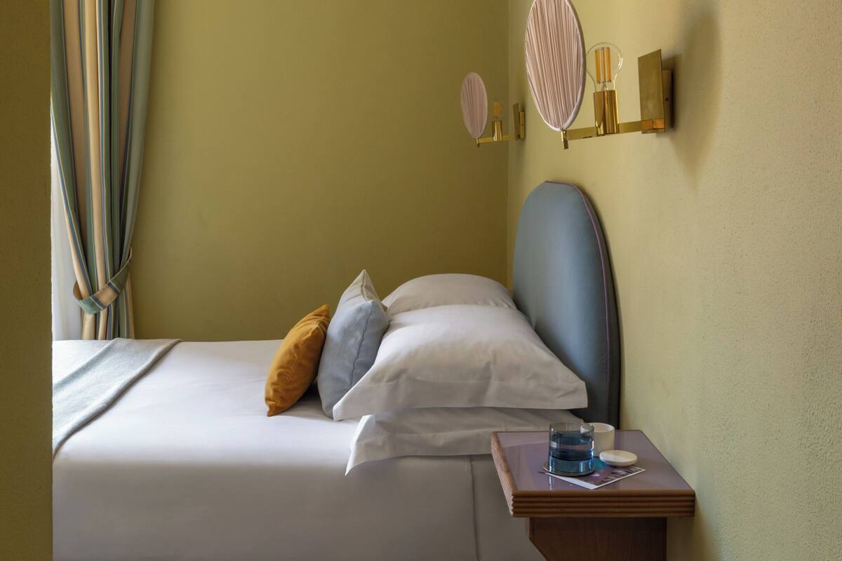 Ein Schlafzimmer mit einem Bett, einem Nachttisch, einem wandmontierten Spiegel und einem Fenster mit Vorhängen.