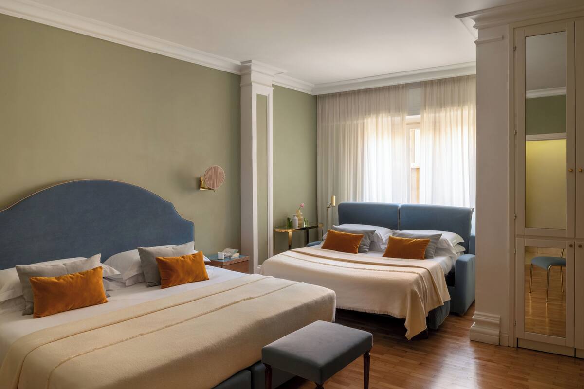 Ein Hotelzimmer mit zwei Betten, einer Bank, einem Nachttisch, einer Lampe und einem fensterbepflanzten Fenster.