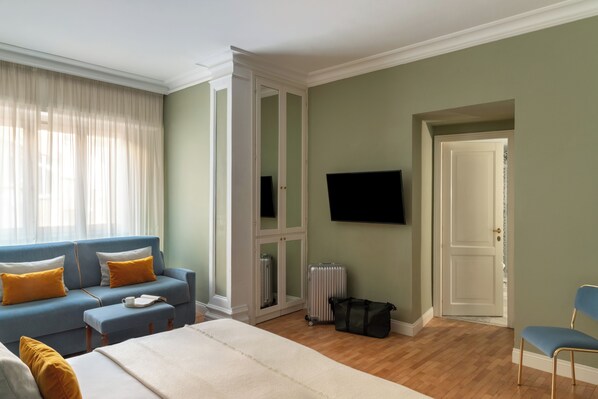 Una habitación con una cama, un sofá azul con cojines amarillos, un televisor en la pared y una puerta que da a otra habitación.