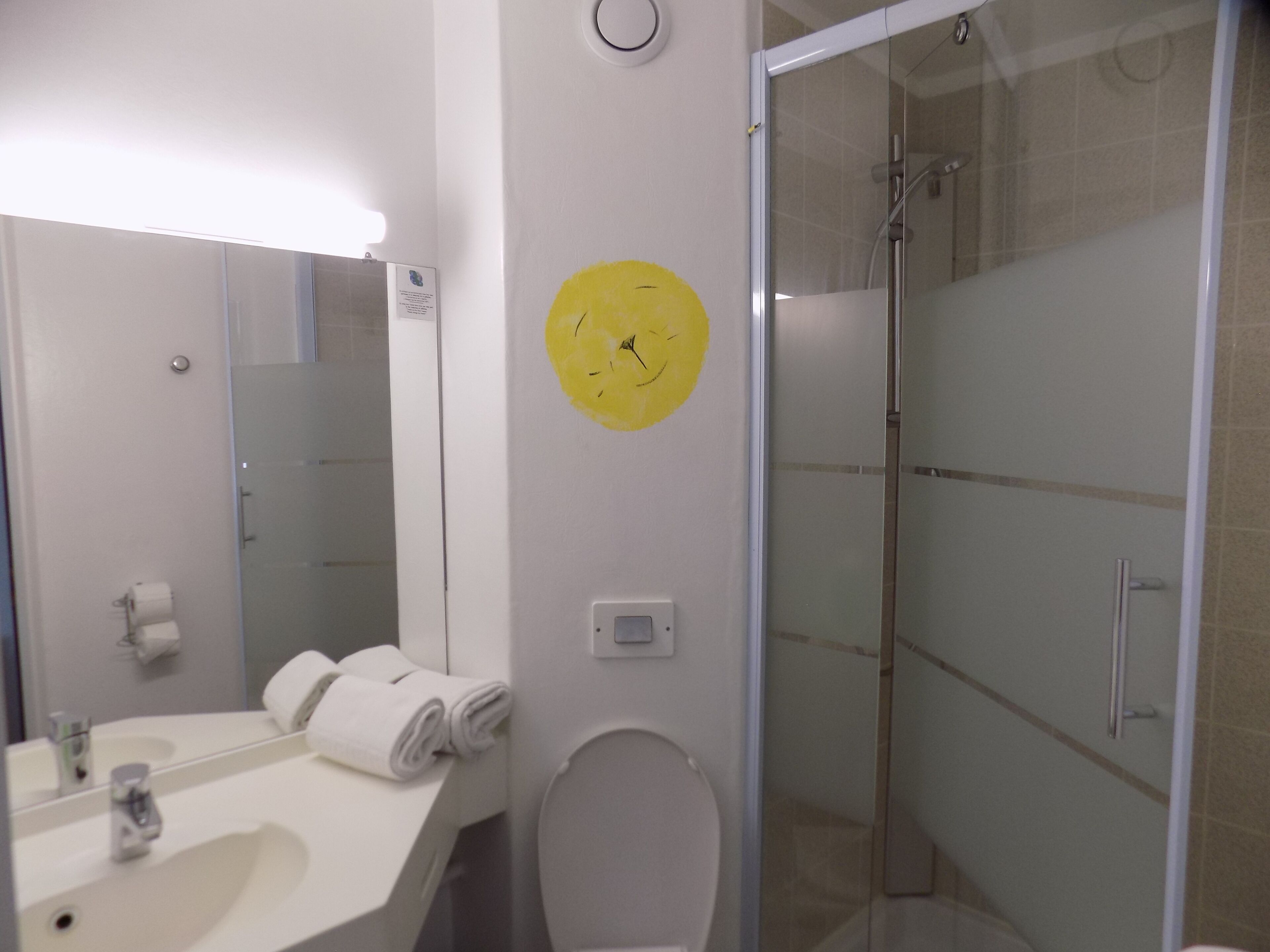 Un baño con lavamanos blanco, espejo y retrete. Hay un reloj amarillo en la pared y una puerta de ducha de vidrio.