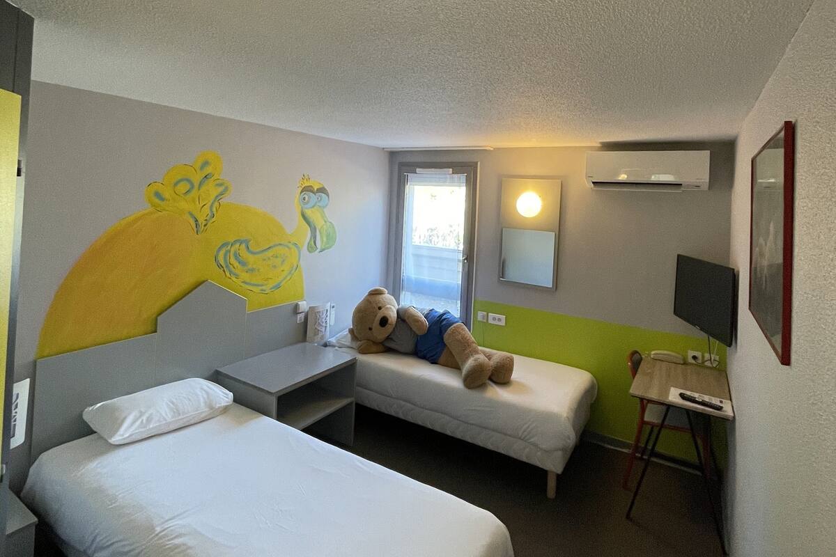 Quarto de hotel com duas camas de solteiro, uma pequena escrivaninha, uma cadeira, uma TV e uma parede com um mural amarelo.
