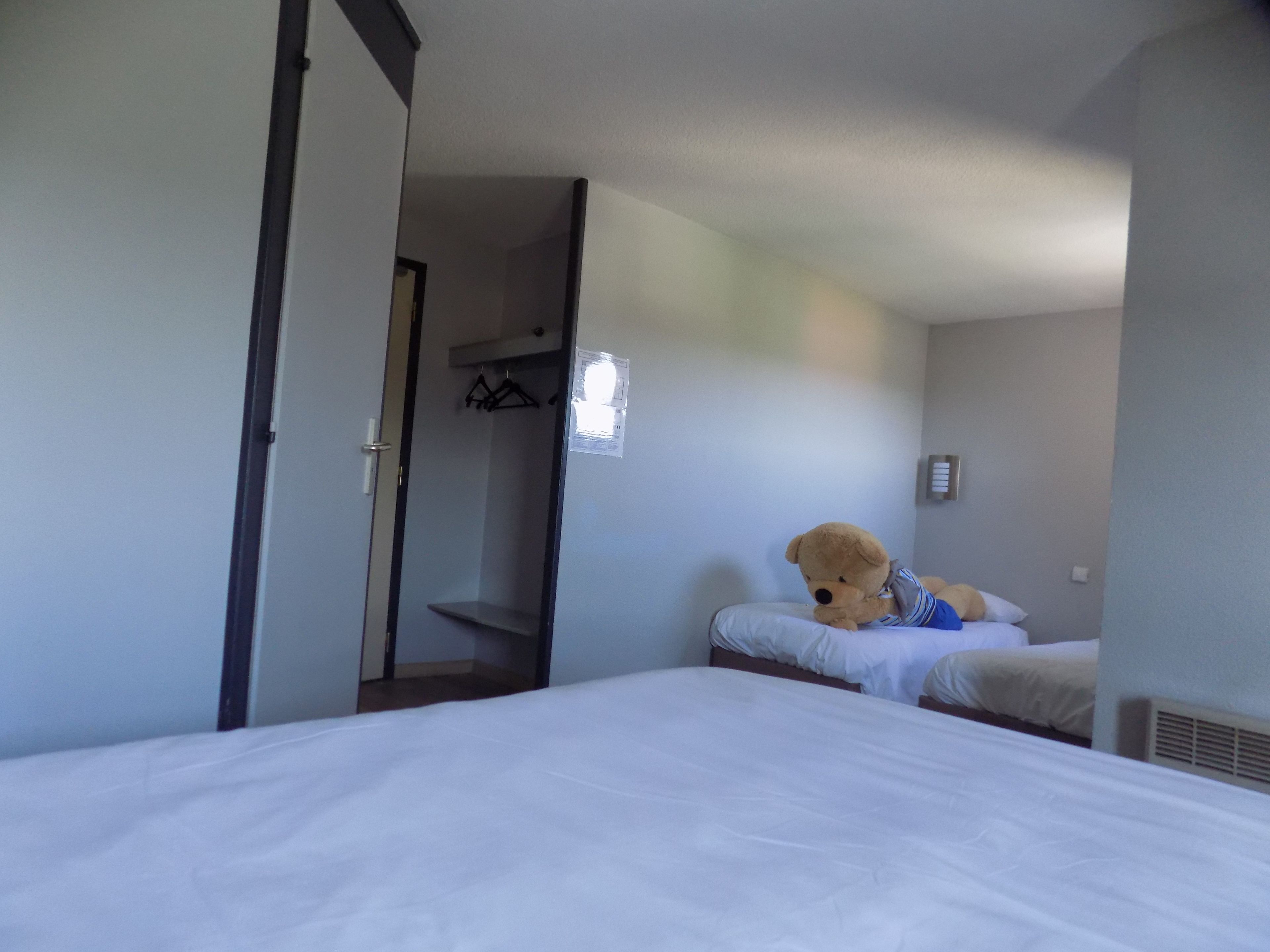 A hotel room with two beds, a teddy bear on one bed, and a coat rack in the corner.