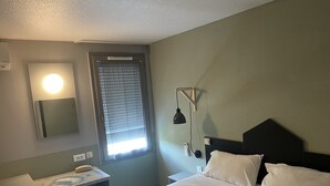 Une chambre d’hôtel avec un lit, une table de chevet, une lampe, un miroir et une fenêtre avec des stores.