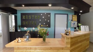 Un comptoir d’accueil en bois, une enseigne indiquant « HÔTEL NESTOR » et un panneau d’accueil. On peut y voir des objets décoratifs, comme une étoile de mer, un vase avec des fleurs et une figurine de girafe.