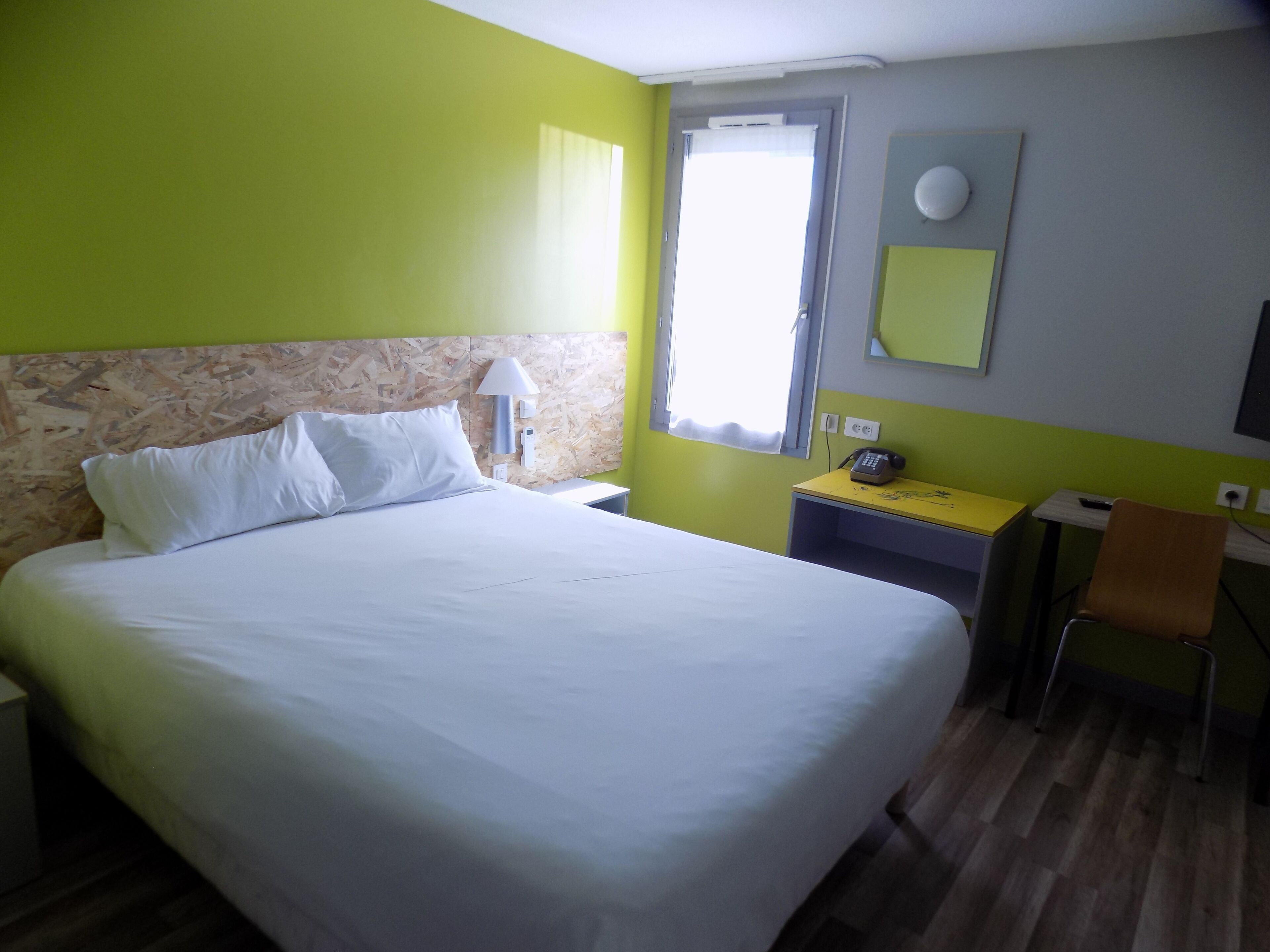 Een hotelkamer met een bed, een bureau met stoel, een lamp, een telefoon en een raam met gordijnen.