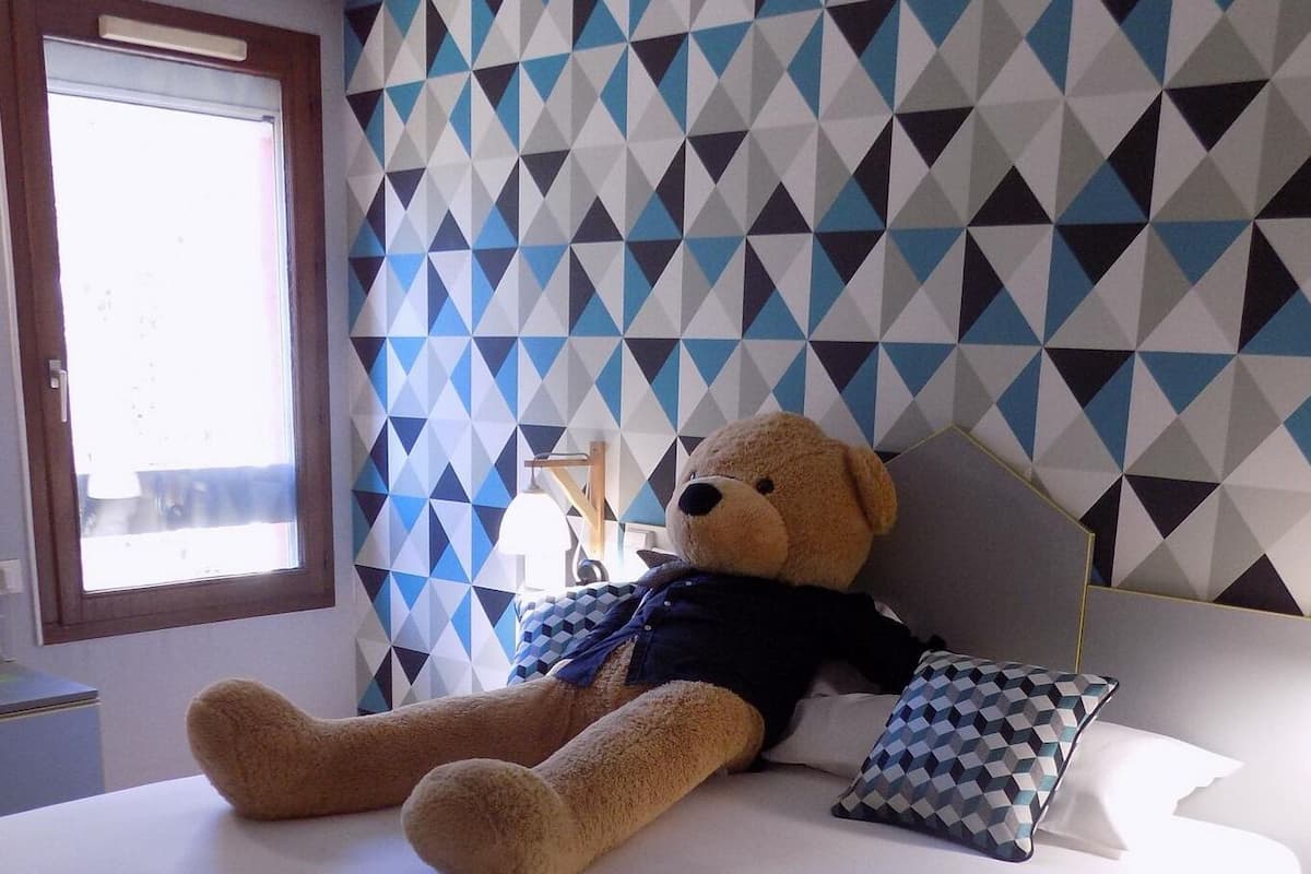 Quarto com uma cama grande, um ursinho de pelúcia, parede com padrão geométrico, uma janela e uma mesa de cabeceira com abajur.