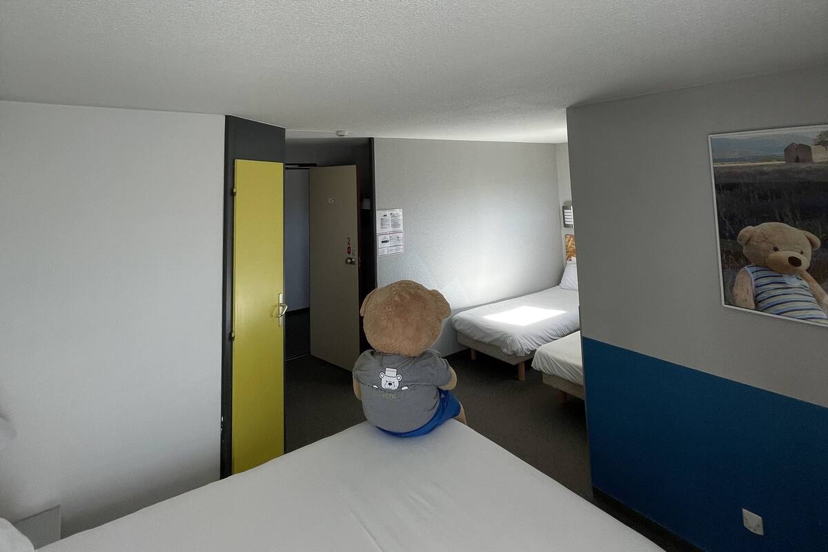 Quarto de hotel com cama, uma pequena escrivaninha, uma cadeira, um ursinho de pelúcia e uma porta que dá para outro cômodo.
