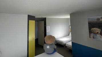 A hotel room with a bed, a small desk, a chair, a teddy bear, and a door leading to another room.