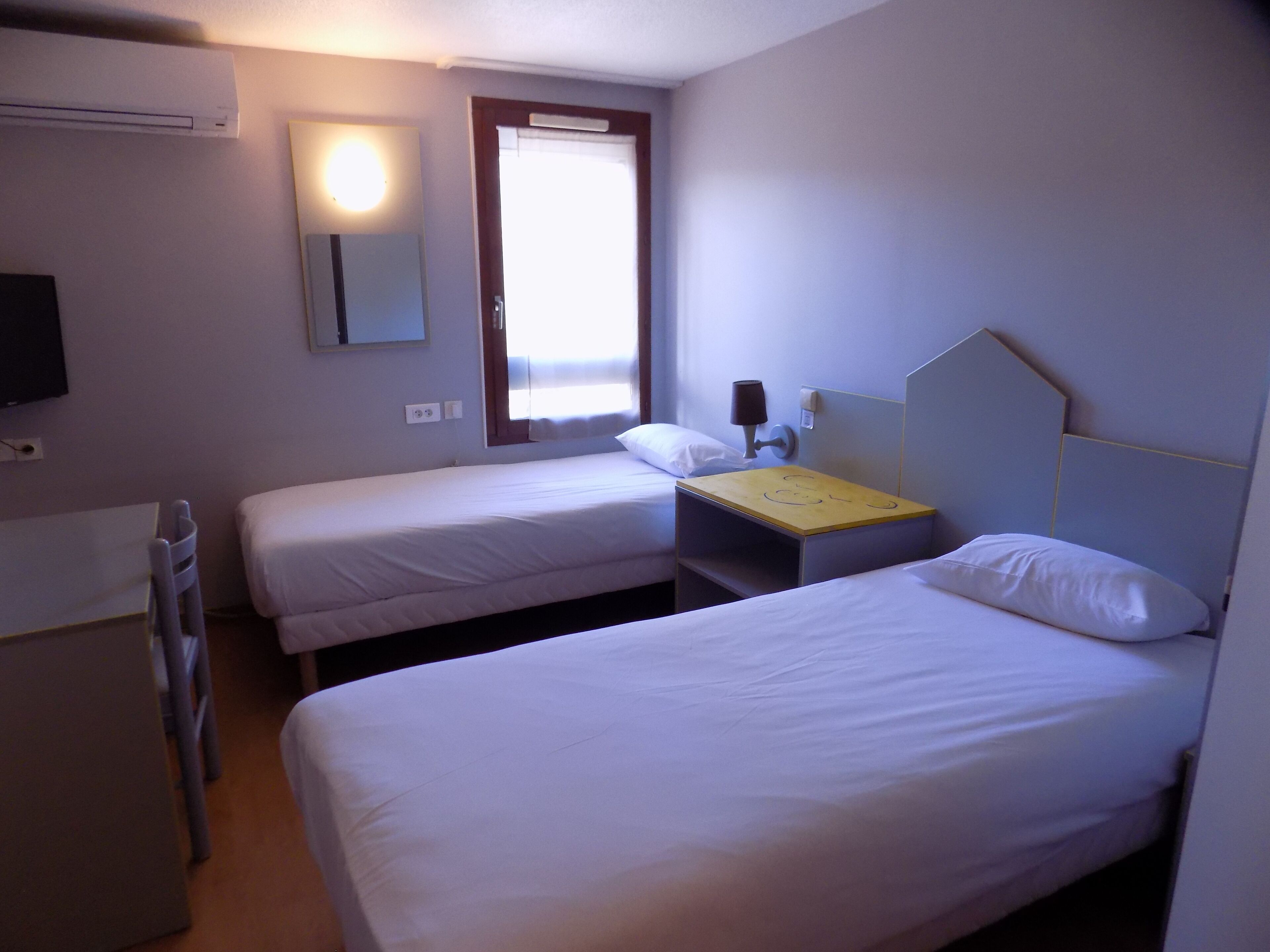 A hotel room with two single beds, a desk, a chair, a TV, and a window with curtains.