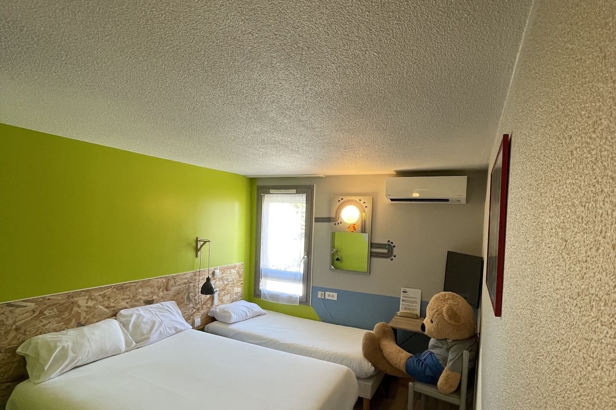 Quarto de hotel com duas camas, uma parede de destaque verde, janela com vista, escrivaninha com cadeira e um urso de pelúcia.