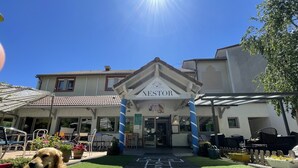 L’entrée d’un hôtel, avec une enseigne portant le nom « NESTOR », des tables et des chaises à l’extérieur, et un chien allongé sur l’herbe.