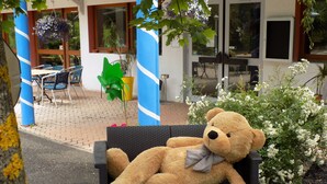 Un ours en peluche est assis sur une chaise en osier dans un espace extérieur aménagé avec des plantes en pot et une table de patio.