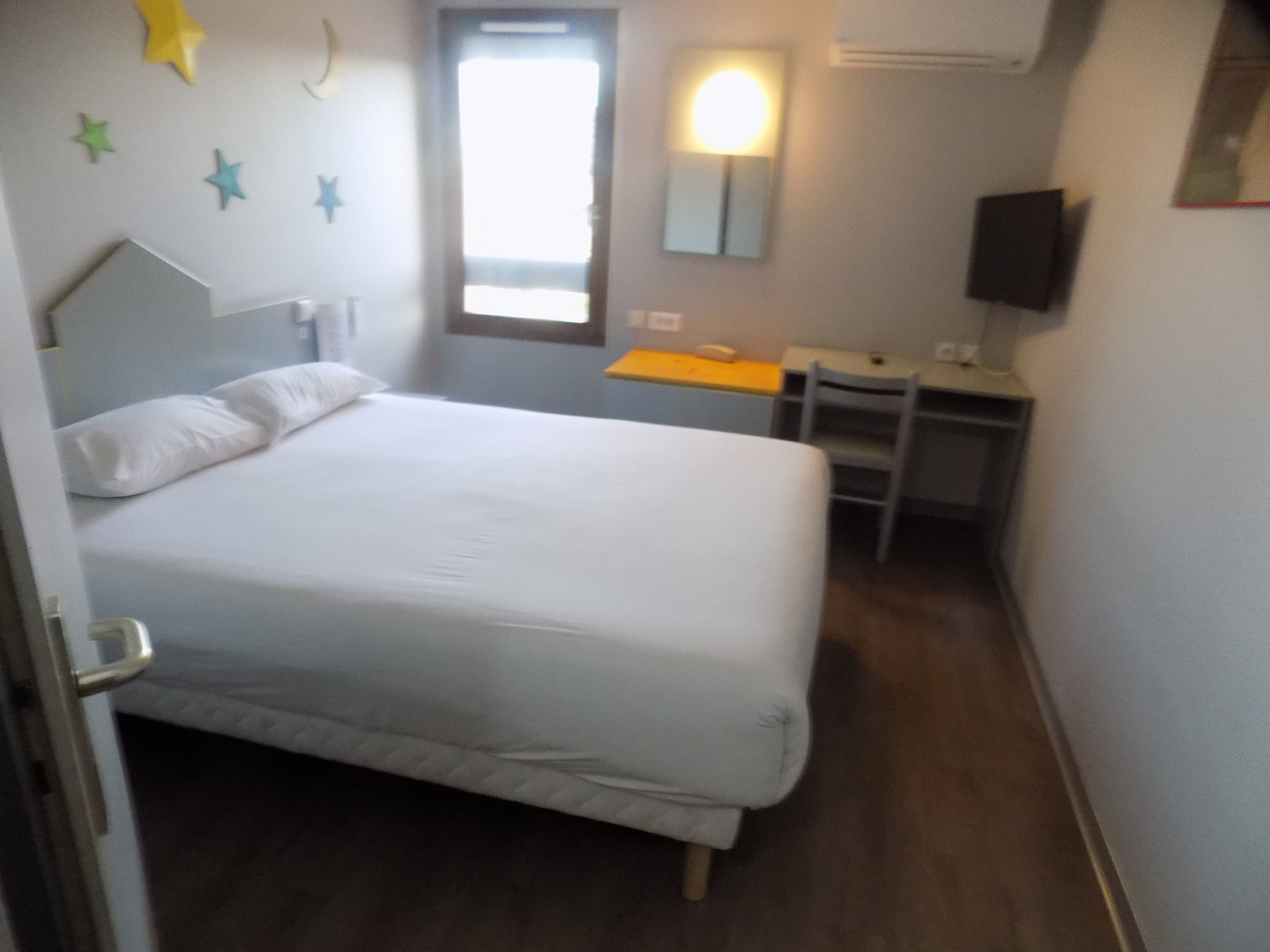 A bedroom with a bed, a desk, and a chair. There is a window with a view of the outside.