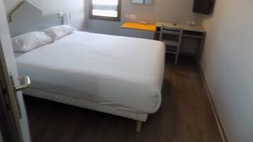 A bedroom with a bed, a desk, and a chair. There is a window with a view of the outside.